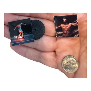May include: Miniature album art and a black vinyl record are held in a hand. The album art features a person skateboarding. Another square image shows a shirtless person. A dime is also in the hand.