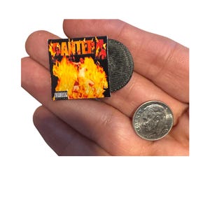 May include: Miniature album cover and vinyl record with the word "PANTERA" in red letters against a fiery background. The album cover has an advisory warning. A dime is next to the album.