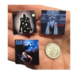 K&ouml;nnte beinhalten: Miniatur-Albumcover mit Alben von Ozzy Osbourne. Die Cover zeigen "Ozzmosis", "Ozzy" und "Bark at the Moon". Eine 10-Cent-M&uuml;nze dient als Gr&ouml;&szlig;envergleich. Die Albumcover sind quadratisch und haben dunkle und farbenfrohe Designs.