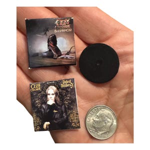 K&ouml;nnte beinhalten: Miniatur-Albumcover von Ozzy Osbournes Alben "Blizzard of Ozz" und "Patient Number 9". Eine schwarze Scheibe und eine M&uuml;nze sind ebenfalls vorhanden. Die Albumcover zeigen Albumkunst und Text. Die M&uuml;nze dient zum Gr&ouml;&szlig;envergleich.