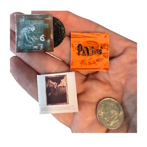 May include: Miniature album covers and a vinyl record are held in a hand. The album covers are for the bands Pixies and Nirvana. One cover has a monkey graphic and the number 6. A dime is also in the hand.