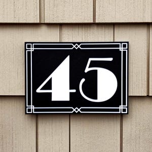May include: A rectangular black house number sign with the number 45 in white. The sign has a white decorative border and is mounted on a light brown shingle siding.