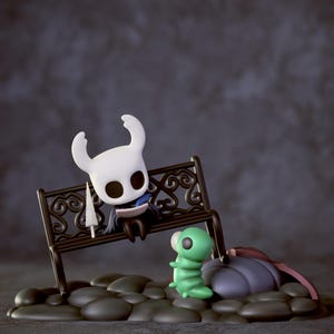 May include: A figurine scene featuring characters from Hollow Knight. A white character with a large head sits on a decorative bench, holding a sword. A green caterpillar-like creature stands nearby. The scene is set on a stone base.