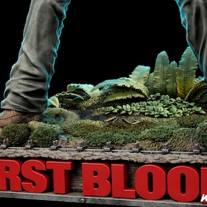 以下が含まれることがあります： 太い赤文字でFIRST BLOODと書かれた詳細なディスプレイベース。ベースは緑のシダ、苔、散らばった金属製の薬莢がある森の地面を表現している。茶色のズボンと茶色のブーツが質感のある地面の上に立っている。