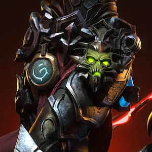 May include: A dark metallic armor piece featuring a central green glowing skull emblem, a circular pendant with a light blue glowing rune, and intricate sculpted details. The armor has a weathered, textured finish with sharp edges and dark tones.