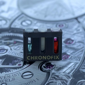 May include: A black rectangular tool with three clear vials, each containing a different colored liquid: blue, white, and red. The word "CHRONOFIX" is printed on the front. The tool rests on a detailed watch mechanism.