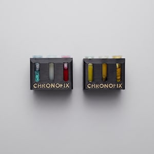 May include: Two black rectangular holders with the text CHRONOFIX printed in gold. Each holder contains three small glass vials. The left holder has blue, white, and red liquids. The right holder contains three vials filled with yellow liquid.