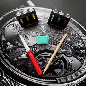Puede incluir: Un surtido de herramientas y suministros para la reparación de relojes, que incluye dos contenedores negros etiquetados "CHRONOFIX" con pequeños viales de líquidos de colores. También están presentes una herramienta roja y blanca, un palillo de madera y un pequeño cuadrado verde, todo sobre una esfera de reloj detallada.