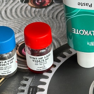 Op de afbeelding: Een assortiment uurwerksmeermiddelen en pasta. Twee kleine glazen flesjes met blauwe en rode doppen zijn gelabeld met "SYNT-A-LUBE" en "SYNT-HP 1300". Een tube "MOLYKOTE DX Paste" is ook zichtbaar. Alle producten zijn van Zwitserse fabrikanten.