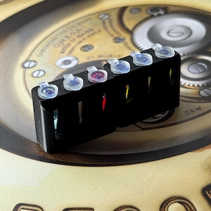 Può includere: Uno strumento nero per la riparazione di orologi con sei contenitori trasparenti contenenti liquidi colorati. Lo strumento è posto su uno sfondo di un quadrante di orologio vintage con ingranaggi e testo visibili.