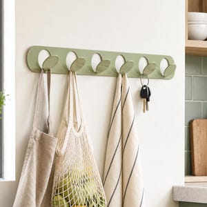May include: A sage green wall-mounted coat rack featuring six circular cutouts with integrated flip-down hooks. The rack holds a beige apron, a cream-colored mesh produce bag filled with green apples and a lemon, a striped kitchen towel, and a set of keys.