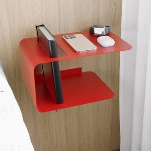 以下が含まれることがあります： モダンな赤い壁掛け式ナイトスタンド。2段の棚があり、上段にはスマートフォン、スマートウォッチ、マウスが置かれています。下段には本とタブレットが置かれています。明るい木製のパネルの壁に取り付けられています。