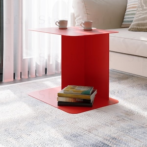 Puede incluir: Una mesa auxiliar roja brillante con una parte superior rectangular y una sola pata ancha. Dos tazas pequeñas de color claro están sobre la mesa. Una pila de libros descansa sobre el estante inferior. La mesa está junto a un sofá blanco y una ventana con cortinas transparentes.