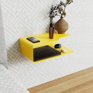 Może przedstawiać: Jasnożółta półka ścienna z dwoma poziomami, zamontowana na wzorzystej białej ścianie. Na górnej półce znajduje się brązowy wazon z suszonymi roślinami, mała brązowa butelka i smartfon. Na dolnej półce znajduje się czarny tablet, smartwatch i biała mysz.