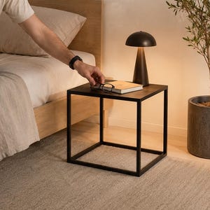Könnte beinhalten: Ein würfelförmiger Beistelltisch aus schwarzem Metall mit schlankem Rahmendesign. Auf der Tischplatte steht eine kleine, dunkelbraune pilzförmige Lampe, die ein warmes Licht nach unten abgibt, sowie ein geschlossenes Buch und eine schwarze Brille.