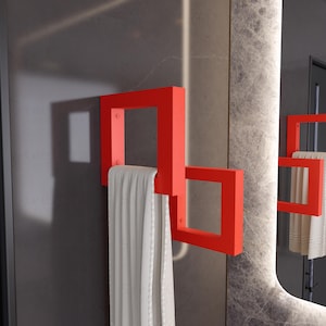 May include: A modern, geometric towel rack in a vibrant red hue. The rack features a square design with a towel draped over it. The rack is mounted on a gray wall, with a mirror in the background. The towel is a light beige color.