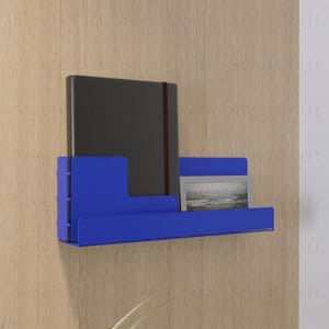 May include: A contemporary blue metal wall shelf, holding a dark grey notebook and a small framed picture. The shelf is mounted on a light wood-effect wall. The design is minimalist.
