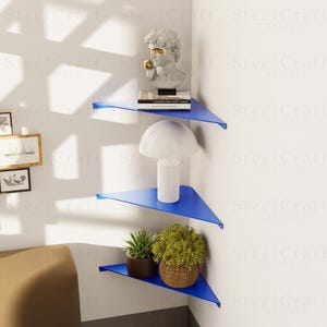 May include: Three bright blue corner shelves fixed to a white wall. The triangular shelves display a bust, a lamp, and potted plants. The shelves are made of metal. The image shows a modern interior design.