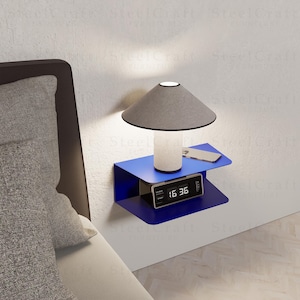 Puede incluir: Una mesita de noche azul moderna montada en la pared con un estante incorporado. Cuenta con una lámpara gris con base blanca, un reloj digital que muestra "16:36" y un teléfono inteligente. La mesita está montada en una pared blanca.