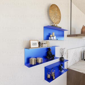 Op de afbeelding: Een modern interieur met drie helderblauwe wandplanken. De planken zijn versierd met decoratieve items, waaronder kaarsen, sculpturen en ingelijste kunstwerken. Boven de bovenste plank is een groot, goudkleurig, cirkelvormig kunstwerk gemonteerd.