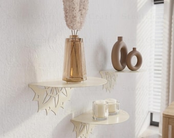Puede incluir: Tres estantes de pared de metal color crema con recortes decorativos de hojas. El estante superior contiene un jarrón de vidrio marrón con flores secas. El estante del medio muestra dos jarrones de cerámica marrones. El estante inferior tiene dos candelabros de vidrio con velas encendidas.