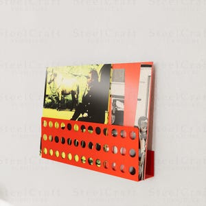 May include: A wall-mounted, bright red magazine rack with a perforated design. The rack holds several magazines and papers with various images. The rack is made of metal and is mounted on a white wall.