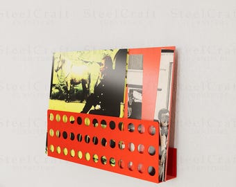 Puede incluir: Un revistero rojo brillante montado en la pared con un diseño perforado. El revistero contiene varias revistas y papeles con varias imágenes. El revistero es de metal y está montado en una pared blanca.