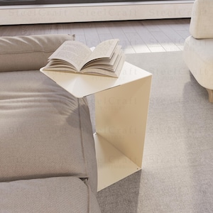 Könnte beinhalten: Ein moderner, weißer, geometrischer Beistelltisch mit einem offenen Buch. Der Tisch hat ein einzigartiges, eckiges Design und steht neben einem grauen Sofa. Der Tisch ist aus Metall und ein zeitgenössisches Möbelstück.
