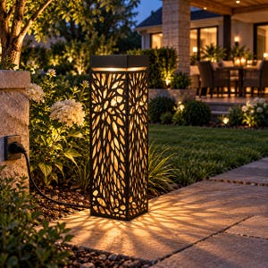 May include: A tall, rectangular outdoor lantern with a black metal frame featuring a cut-out leaf pattern. The lantern emits a warm, glowing light from within, casting intricate shadows on the stone patio surface. A black power cord is visible.