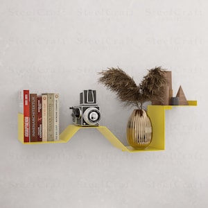 Könnte beinhalten: Modernes gelbes Metall-Wandregal mit einem einzigartigen Wellendesign. Das Regal zeigt Bücher, eine Vintage-Kamera, eine goldene Vase mit getrockneten Pflanzen und dekorative Holzformen. Das Regal ist an einer weißen Wand befestigt.