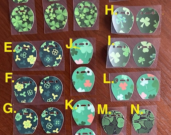 May include: A set of 14 green shamrock patterned stickers for Airpods. The stickers are in various designs, including solid green shamrocks, shamrocks with white outlines, and shamrocks with white dots. The stickers are labeled with letters A through N.