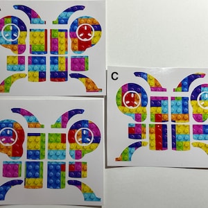 May include: Three white rectangular sheets labeled A, B, and C, each featuring a butterfly-shaped graphic composed of multicolored interlocking plastic brick patterns. The butterfly designs include circular white icons on the upper wings.