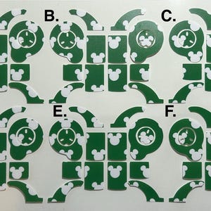 May include: Green die-cut stickers with white Mickey Mouse head shapes and circular designs. The stickers are arranged in a grid pattern, with various shapes and sizes. The overall theme is related to Disney.