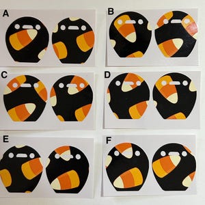 May include: A collection of six pairs of Halloween-themed stickers. Each sticker features a black circular design with candy corn accents in orange, yellow, and white. The stickers have a cartoonish face.