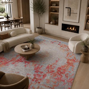 Peut inclure: Un tapis rectangulaire présentant un motif abstrait avec des taches rouge vif et orange superposées sur un fond gris clair et bleu, recouvrant le sol d'un salon avec un canapé, des fauteuils et une table basse ronde en bois.