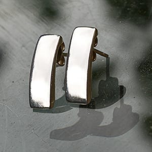Puede incluir: Un par de pendientes de barra curvada de color plateado. Los pendientes tienen una superficie pulida y reflectante y un cierre de poste y embrague. Los pendientes est&aacute;n colocados sobre un fondo borroso, con sus sombras visibles.