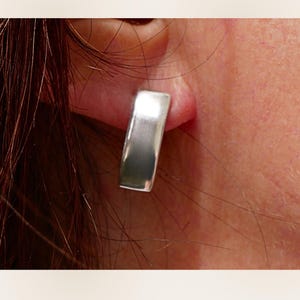 Puede incluir: Primer plano de un pendiente rectangular plateado. El pendiente es de metal macizo pulido con una forma ligeramente curvada. El pendiente se lleva en la oreja de una persona, con la piel visible en el fondo.