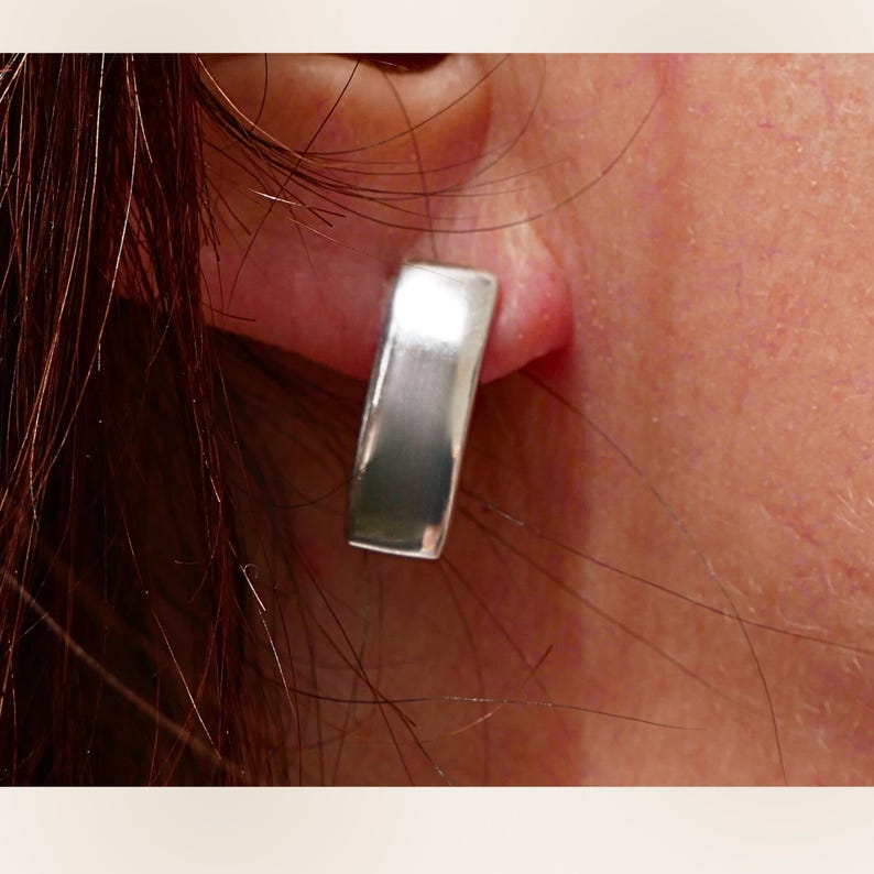 Puede incluir: Primer plano de un pendiente rectangular plateado. El pendiente es de metal macizo pulido con una forma ligeramente curvada. El pendiente se lleva en la oreja de una persona, con la piel visible en el fondo.