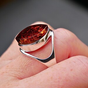 Peut inclure: Bague en argent ornée d'une grande pierre d'ambre ovale. L'ambre est d'un brun rougeâtre profond avec des motifs internes. La bague a un design de bande ouvert et moderne.