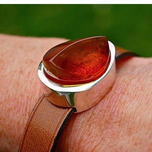 May include: A bracelet with a large, teardrop-shaped amber stone set in a silver bezel. The bracelet has a brown leather strap. The amber stone is a rich, translucent orange-brown colour. The leather strap is brown.