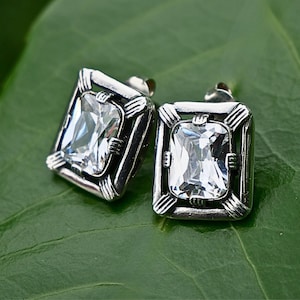 Peut inclure: Une paire de boucles d'oreilles carrées en argent. Chaque boucle d'oreille présente une grosse pierre rectangulaire transparente sertie dans un cadre décoratif. Les boucles d'oreilles sont posées sur une feuille verte.