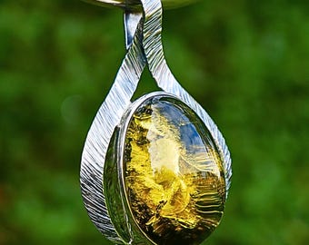 May include: A silver pendant necklace with a large, oval-shaped amber stone. The amber has a golden-yellow hue with internal patterns. The pendant is suspended from a silver loop. The background is a blurred green.
