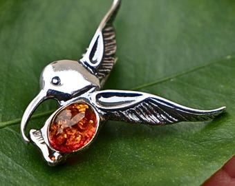 May include: A silver hummingbird pendant with an amber-colored gemstone in its center. The pendant is detailed with etched wings and a long beak. The hummingbird is positioned on a green leaf, showcasing the pendant's design and color.