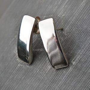 Puede incluir: Un par de pendientes de plata pulida. Cada pendiente tiene forma rectangular con una ligera curva. Los pendientes se muestran sobre una superficie gris, reflejando la luz y creando reflejos.