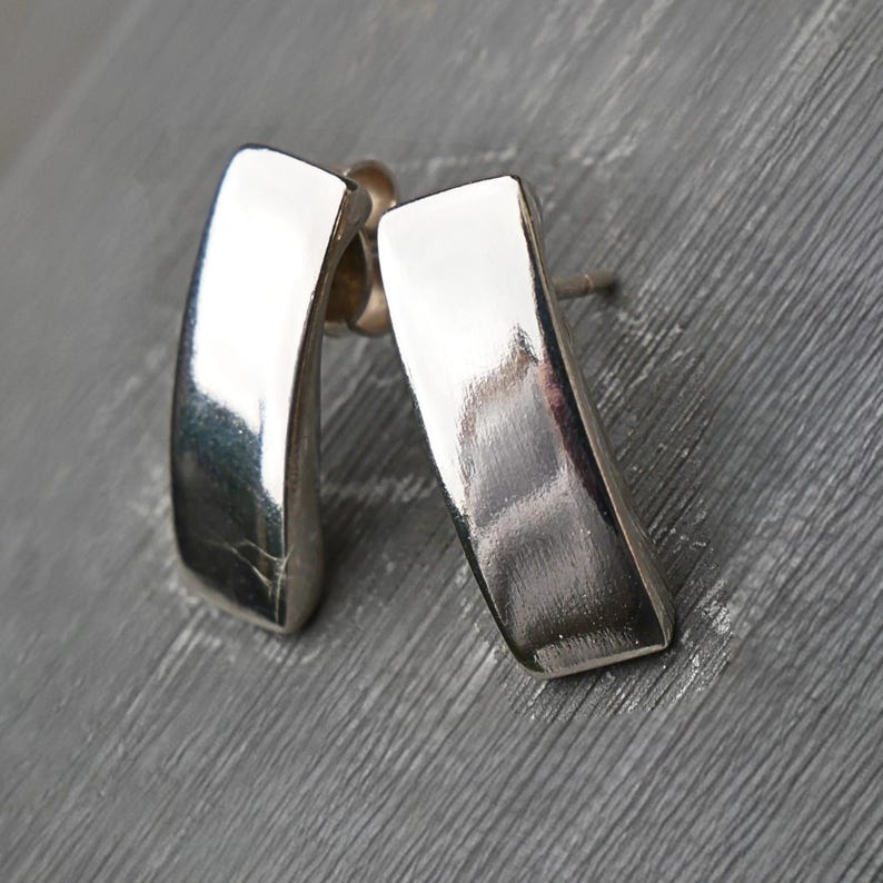 Puede incluir: Un par de pendientes de plata pulida. Cada pendiente tiene forma rectangular con una ligera curva. Los pendientes se muestran sobre una superficie gris, reflejando la luz y creando reflejos.