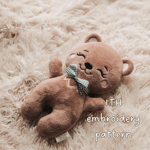 Może przedstawiać: Brązowy pluszowy miś z haftowanymi detalami i niebiesko-białą kratkowaną kokardą. Miś leży na miękkiej, jasnej powierzchni. Tekst na zdjęciu brzmi "ITH embroidery pattern."