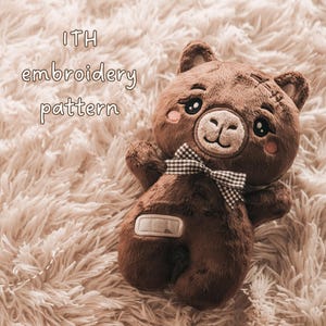 May include: A brown plush bear toy featuring embroidered facial details, pink cheeks, a brown and white gingham bow tie, and a rectangular patch on its belly. The text ITH embroidery pattern is visible in the upper left corner of the image.
