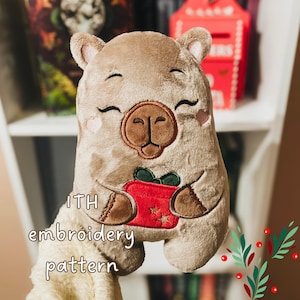 Peut inclure: Une peluche capybara beige avec des traits du visage brodés, des joues roses et un nez marron foncé. Le capybara tient un cadeau rouge avec un nœud vert et des étoiles dorées. Le texte ITH embroidery pattern est écrit sur l'image.