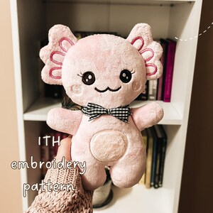 May include: A light pink plush axolotl toy with embroidered black eyes, eyelashes, and a mouth. It features large side gills with pink stitching and a black and white gingham bow around its neck. The text ITH embroidery pattern is visible.