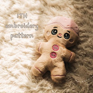 May include: A plush gingerbread person toy with a light pink icing-style head covering, large embroidered eyes, and two pink circular buttons on the chest. The text ITH embroidery pattern is visible on the left side of the plush figure.